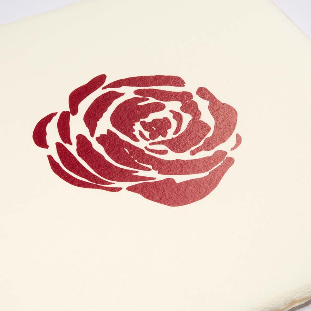 Close-up of a hand-printed red rose motif on textured ivory paper, highlighting the uneven ink and organic paper surface.