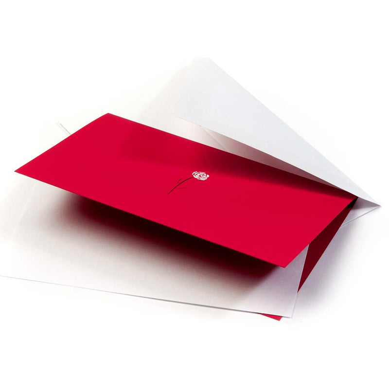 A bright red greeting card resting partly inside a white envelope, shown at a slight angle, with a small white rose and fine black stem printed in the middle.