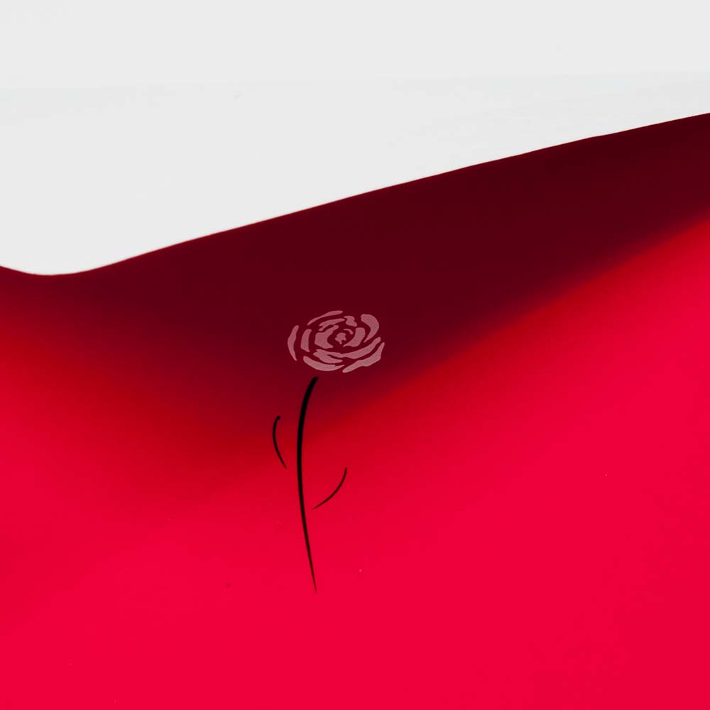 A bright red greeting card shown close up, featuring a small white rose with a thin black stem printed near the center against the smooth red surface.