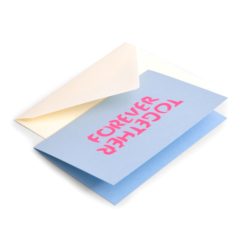 Forever Together greeting card in light blue cardstock, shown partially inside a cream-colored envelope, with bold neon pink brush lettering reading “FOREVER TOGETHER” on the front.