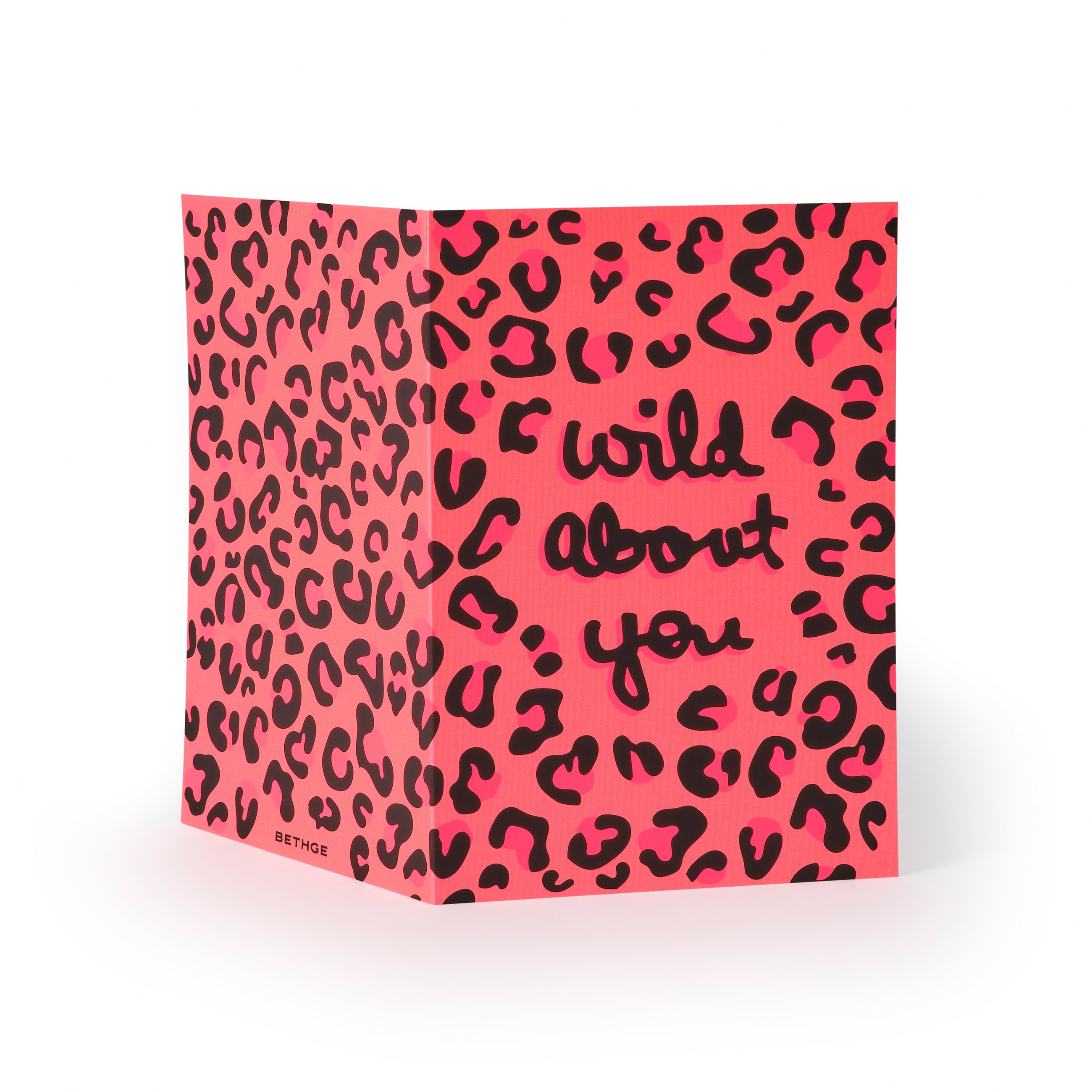 Greeting card opened flat, showing a vibrant pink leopard-print design with expressive black lettering spelling “wild about you” across the front, set against a clean white background.