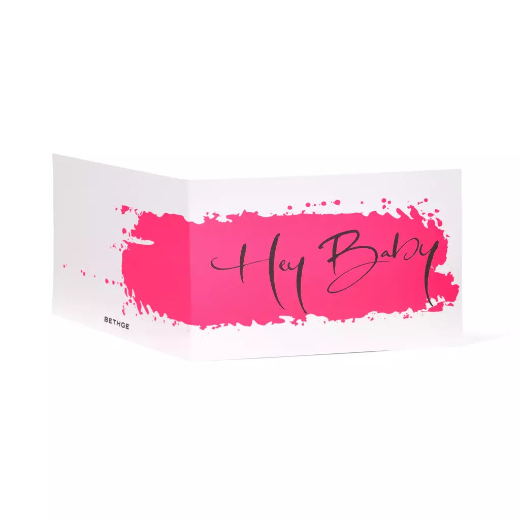 A white folded greeting card shown at an angle, featuring a bold hot-pink paint stroke across the front with the handwritten words “Hey Baby” in flowing black script.