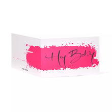 A white folded greeting card shown at an angle, featuring a bold hot-pink paint stroke across the front with the handwritten words “Hey Baby” in flowing black script.