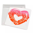 A white greeting card shown slightly angled, featuring a hand-painted heart in a vivid pink-to-orange gradient, filled with flowing handwritten text in white and paired with a matching white envelope.