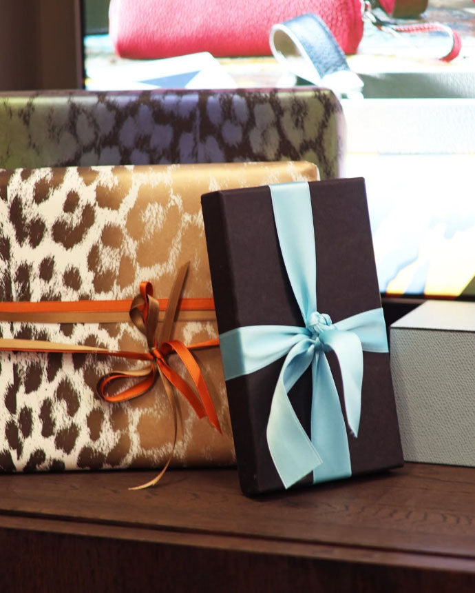 Stacked gift boxes featuring leopard print, solid colors, and satin ribbons in soft blue and warm orange tones, arranged on a wooden surface in natural light.