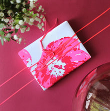 A neatly wrapped gift in white paper with poppies in bright pink and red, tied with thin pink ribbon, set against a rich red background with soft floral accents nearby.