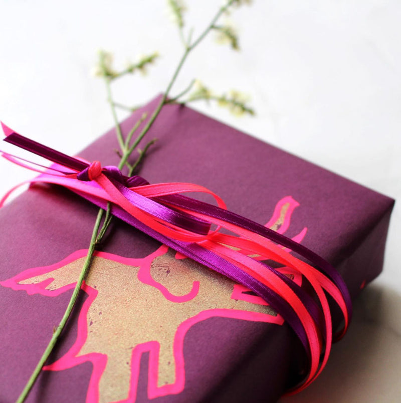 A gift box wrapped in flying elephant wrapping paper, finished with layered pink and magenta ribbons and a delicate sprig of greenery.