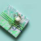 A gift box wrapped in paradise green/gray wrapping paper, tied with layered green and blue ribbons and topped with a white flower, set on a soft teal background.