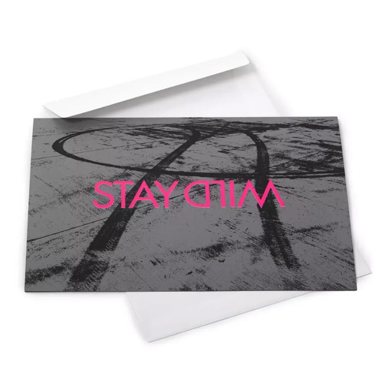 A greeting card placed over a white envelope, showing a gritty black-and-white tire track design with “STAY WILD” printed across it in vivid pink text.