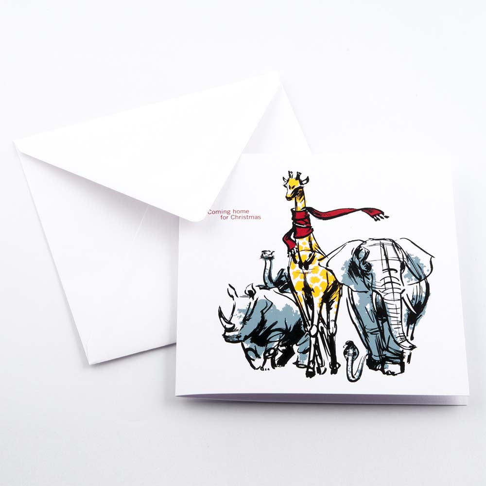 White Christmas card with an illustrated giraffe wearing a red scarf, walking alongside an elephant and a rhinoceros, with the text “Coming home for Christmas," paired with a plain white enevelope.