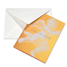 Elegant Christmas card with a shimmering gold-orange background, featuring a large pop-art-style white angel illustration and the text “down to earth.” Includes a white envelope.