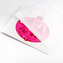 Christmas card with a bright pink ornament illustration filled with silhouettes of festive figures, animals, and decorations, paired with a translucent envelope.