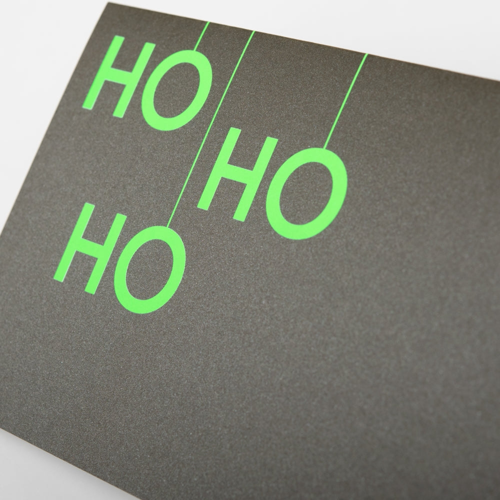 A close-up of a black Christmas card with bold neon green letters arranged vertically to read "HO HO HO".