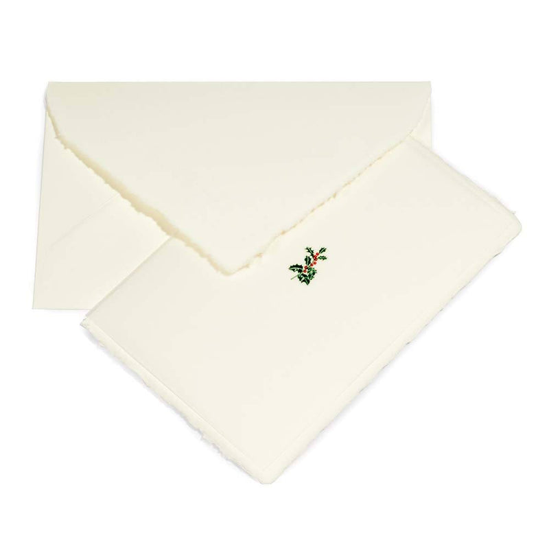 A cream-colored Christmas card with deckled edges, featuring a small holly sprig with green leaves and red berries in the center, shown with a matching cream envelope.