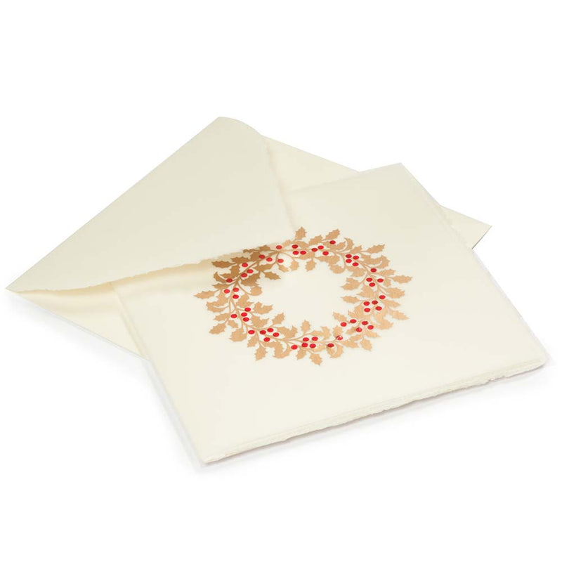 A cream-colored Christmas card with deckled edges, featuring a festive holly wreath design made of gold leaves and bright red berries, shown with a matching cream envelope.