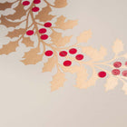 A close-up of a beige Christmas card adorned with a golden holly wreath and red berries.