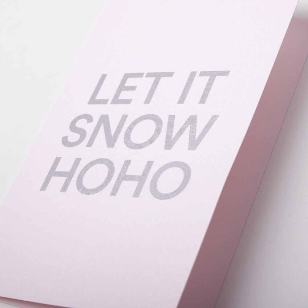 A close-up of a light pink Christmas card with bold silver text reading 'LET IT SNOW HOHO'.