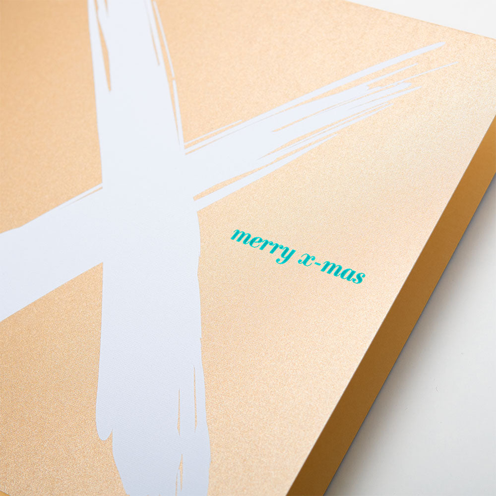 A close-up of a Christmas card with a bold white brushstroke “X” on a gold background and the text “merry x-mas” in small green letters.