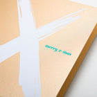 A close-up of a Christmas card with a bold white brushstroke “X” on a gold background and the text “merry x-mas” in small green letters.