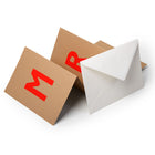 Folded Christmas cards on a white surface, one with the letter “M” and another with the letter “R,” paired with a white envelope.