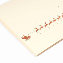 A close-up of a minimalist cream-colored Christmas card showing a row of small gold reindeer pulling a sleigh, numbered up to 31. The reindeer on the 24th is marked in red. Paired with a matching cream envelope.