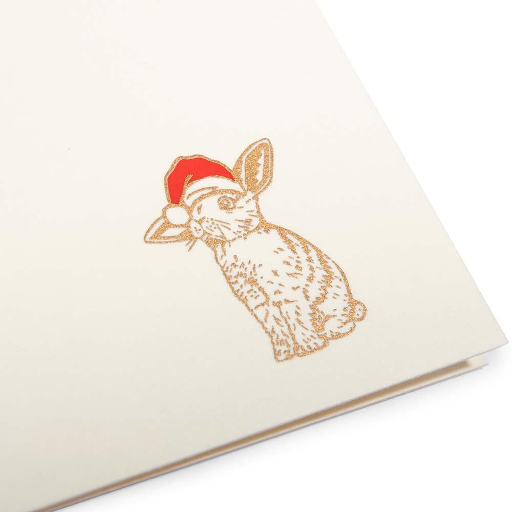 A close-up of a minimalist cream-colored Christmas card with a small illustration of a rabbit wearing a Santa hat in the bottom right corner.