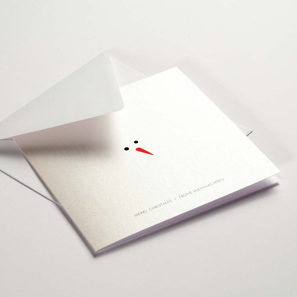 Minimalist Christmas card showing only a snowman's black eyes and orange carrot nose on a shimmering white background, with 'Merry Christmas' and 'Frohe Weihnachten' printed below, paired with a white envelope.