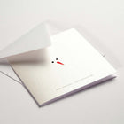 Minimalist Christmas card showing only a snowman's black eyes and orange carrot nose on a shimmering white background, with 'Merry Christmas' and 'Frohe Weihnachten' printed below, paired with a white envelope.