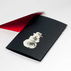 A black Christmas card featuring a rough white sketch of a snowman with a small red carrot nose, paired with a white envelope lined in red.