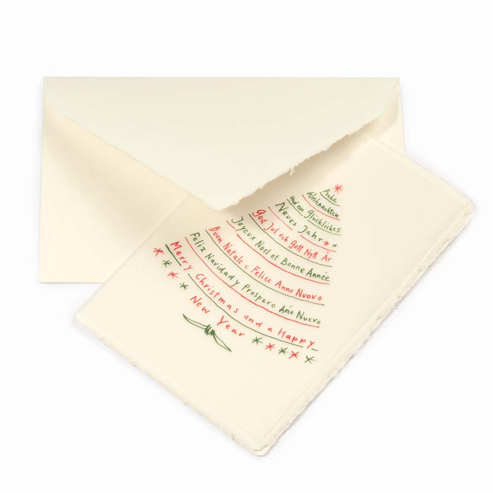 A cream-colored Christmas card with deckled edges, featuring festive greetings in multiple languages written in red and green text arranged in the shape of a Christmas tree, shown with a matching cream envelope.