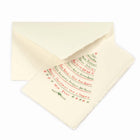 A cream-colored Christmas card with deckled edges, featuring festive greetings in multiple languages written in red and green text arranged in the shape of a Christmas tree, shown with a matching cream envelope.