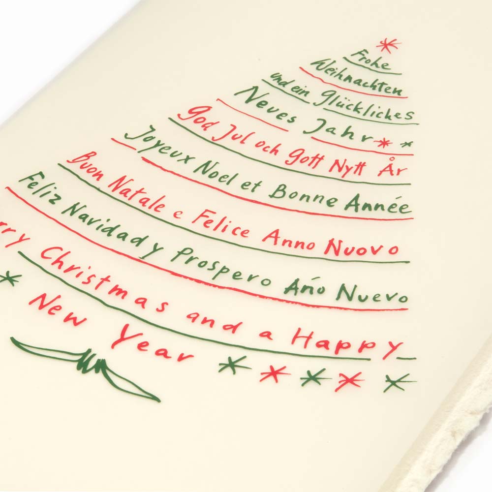 A close-up of a cream-colored Christmas card with deckled edges, featuring festive greetings in multiple languages written in red and green text arranged in the shape of a Christmas tree.