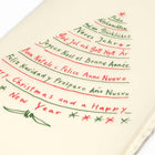 A close-up of a cream-colored Christmas card with deckled edges, featuring festive greetings in multiple languages written in red and green text arranged in the shape of a Christmas tree.
