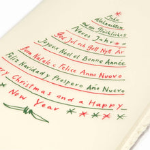 A close-up of a cream-colored Christmas card with deckled edges, featuring festive greetings in multiple languages written in red and green text arranged in the shape of a Christmas tree.