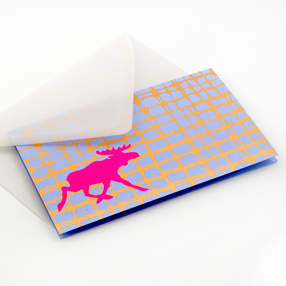 Modern Christmas card with a bright pink moose silhouette against a blue and gold grid-like background, paired with a translucent white envelope.