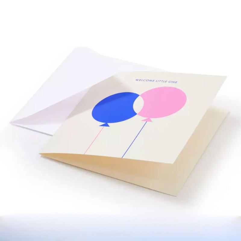 A minimalist greeting card with a soft cream background, featuring two overlapping balloons in blue and pink with fine strings, and the subtle text “WELCOME LITTLE ONE” centered above, tucked into a white envelope.